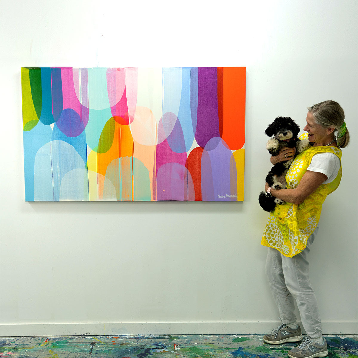 Claire holding her puppy, Iris, standing next to her original painting, Deliberately Divine, hung on her studio wall.